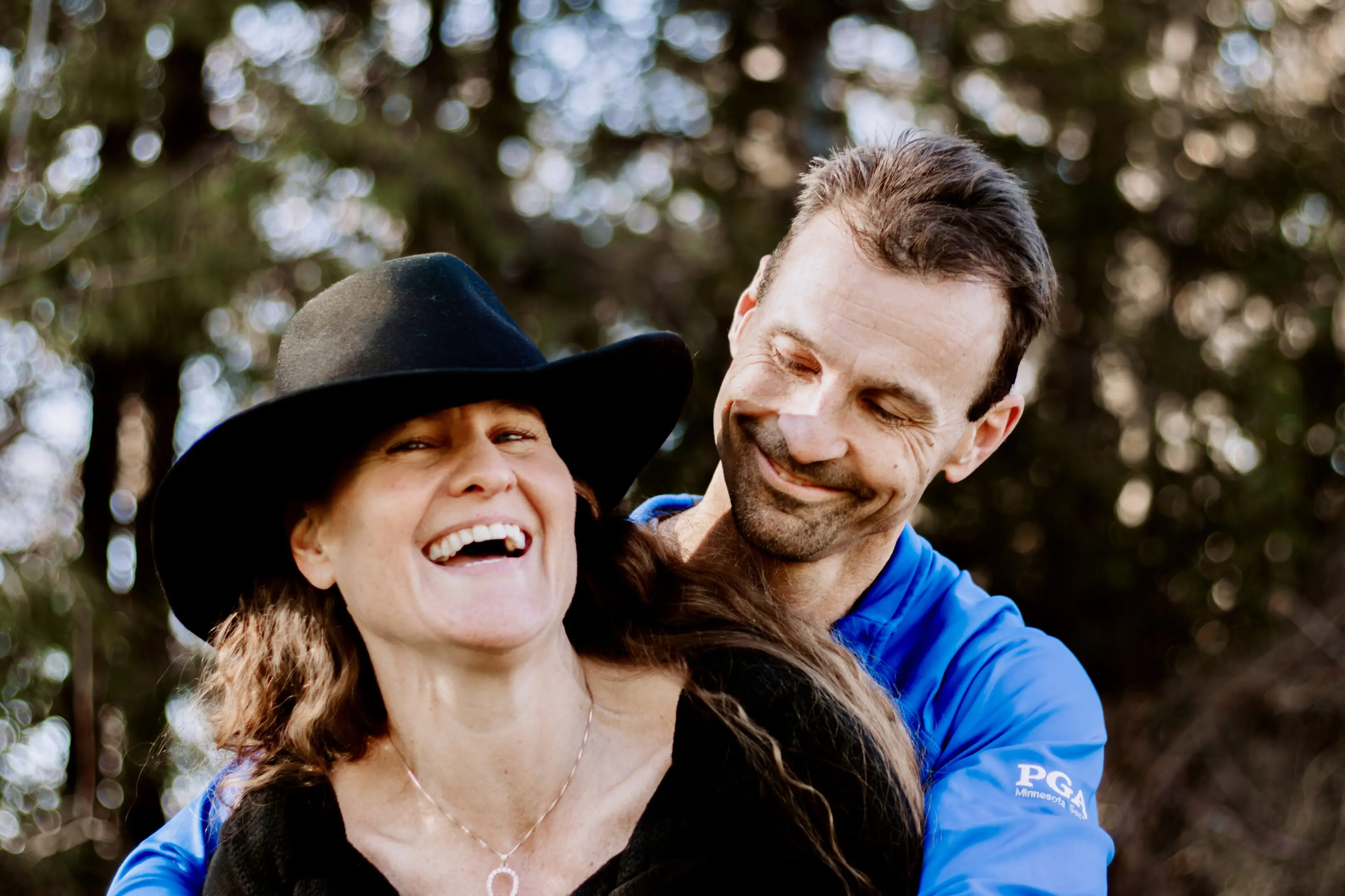 A joyful couple sharing a laugh outdoors, with the man in blue and the woman in a black hat.